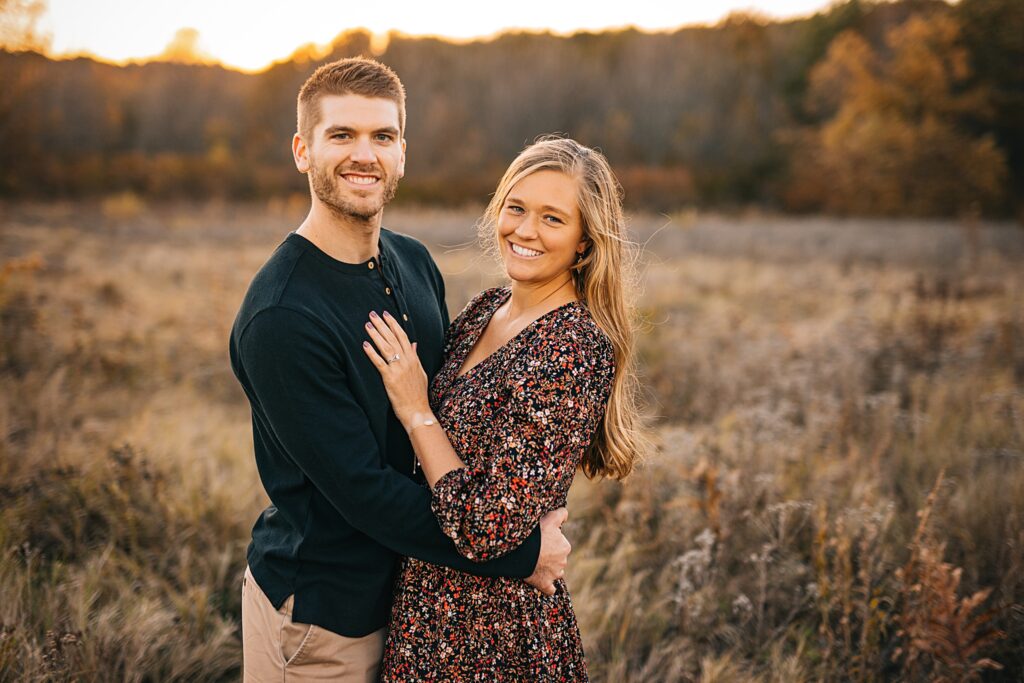 a husband and wife pose for a portrait in a prairie at sunset