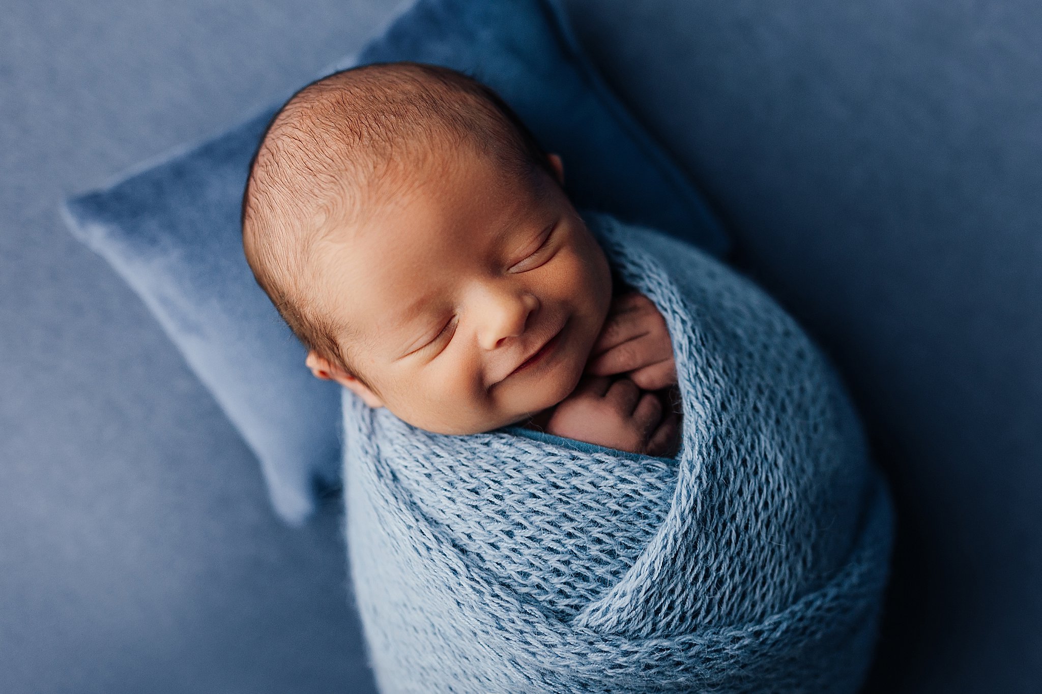 a smiling baby boy wrapped in baby blue knit wrap on a soft blue pillow at a newborn photography session in a studio in cedar falls, iowa